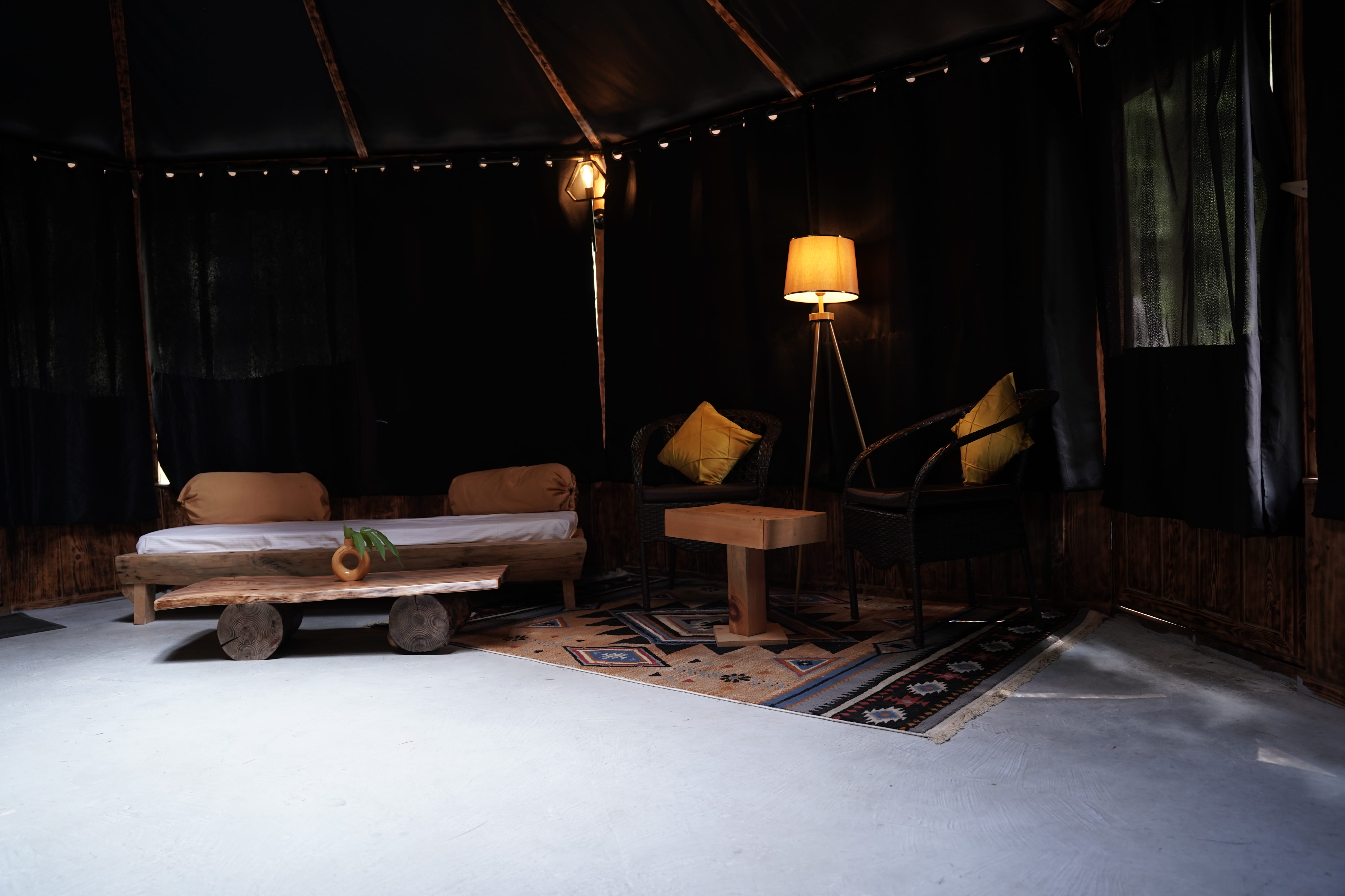 Dark moody lounge with wooden daybed, wicker chairs, tripod floor lamp, and string lights